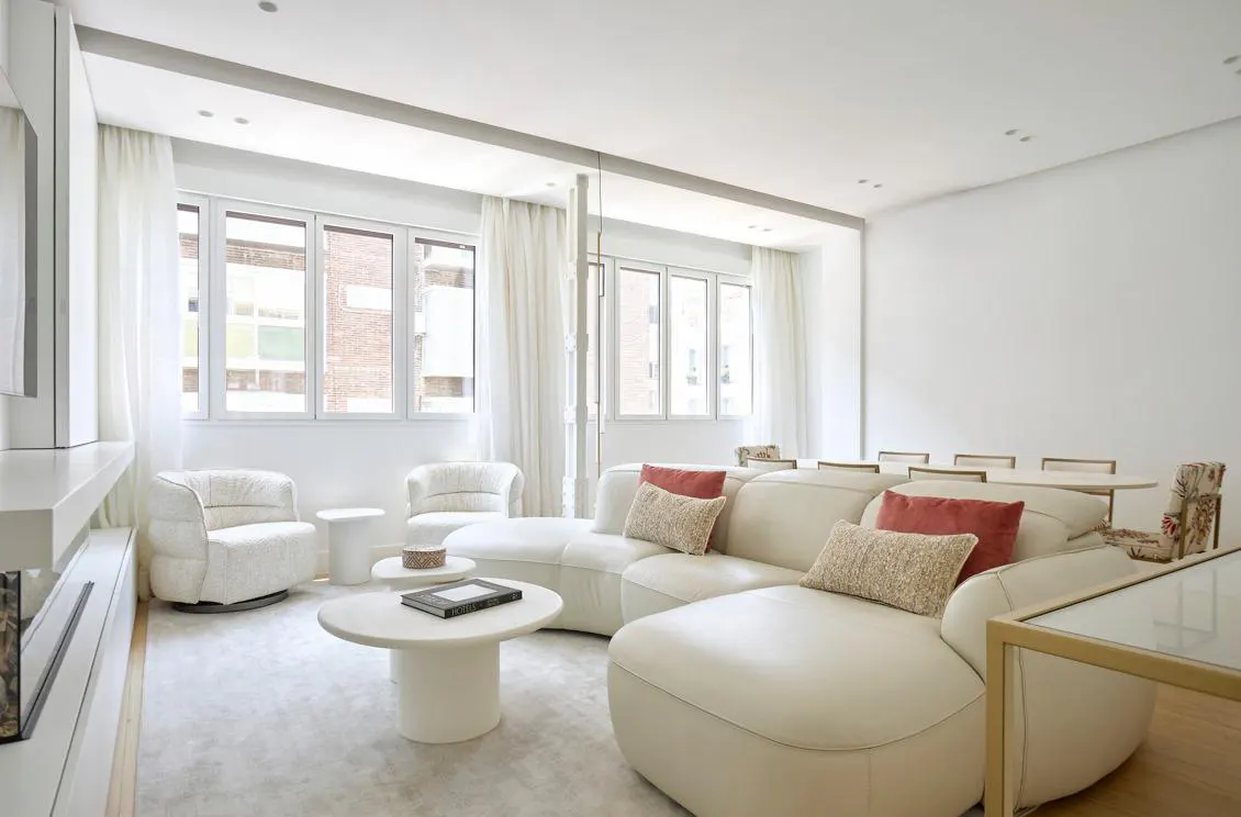 Bright, all-white living room with a curved sofa, round tables, and large windows with sheer curtains. Red and beige pillows add color.