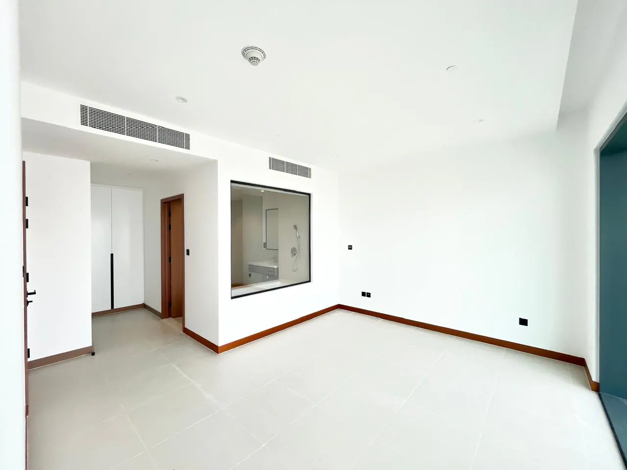 Bright, empty room with white walls, tile floor, and wood trim. A window shows a bathroom with a shower.