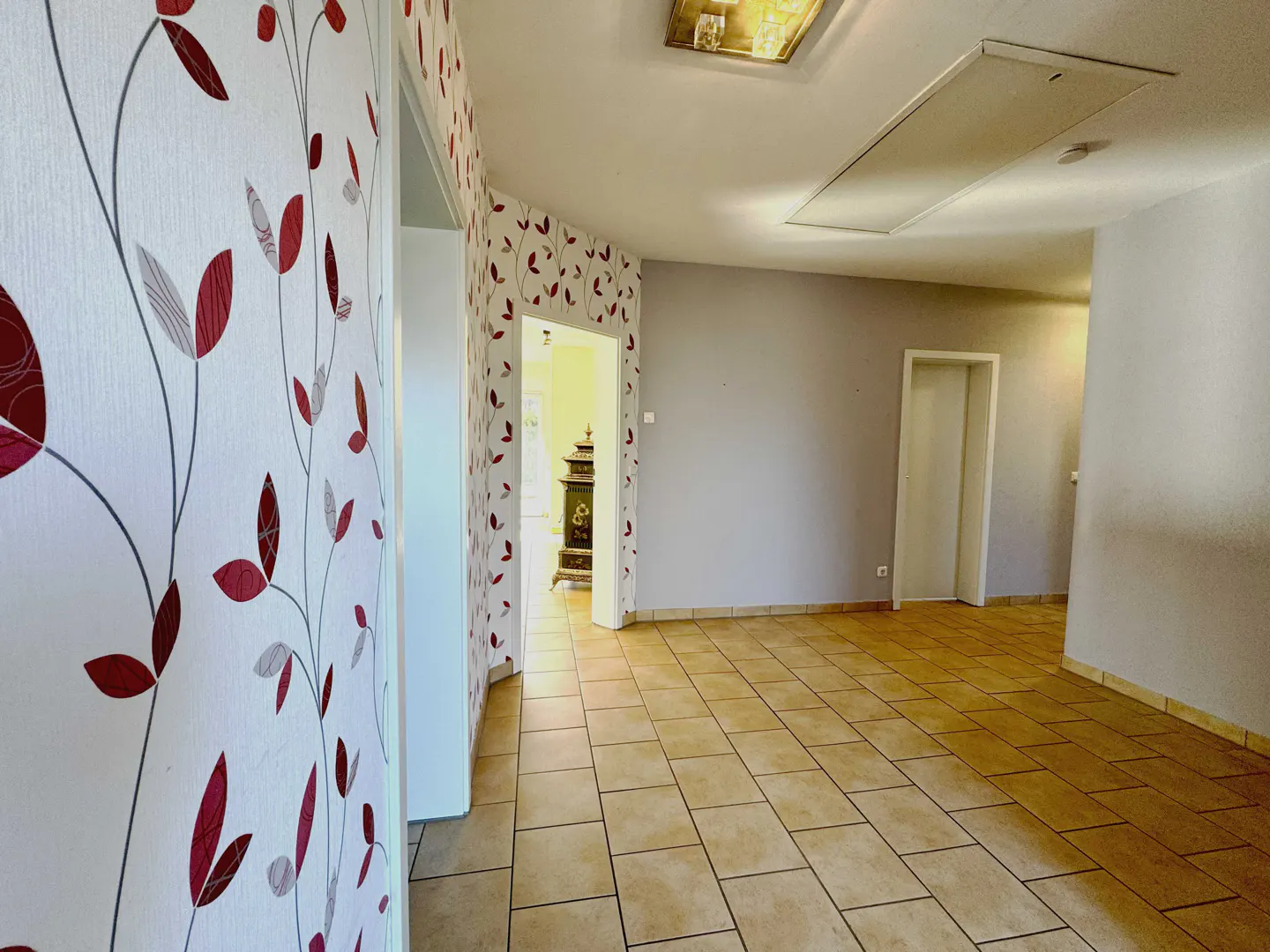 Hallway with beige tile floor, white walls, and red leaf wallpaper. A doorway leads to a room with a dark wood stove.