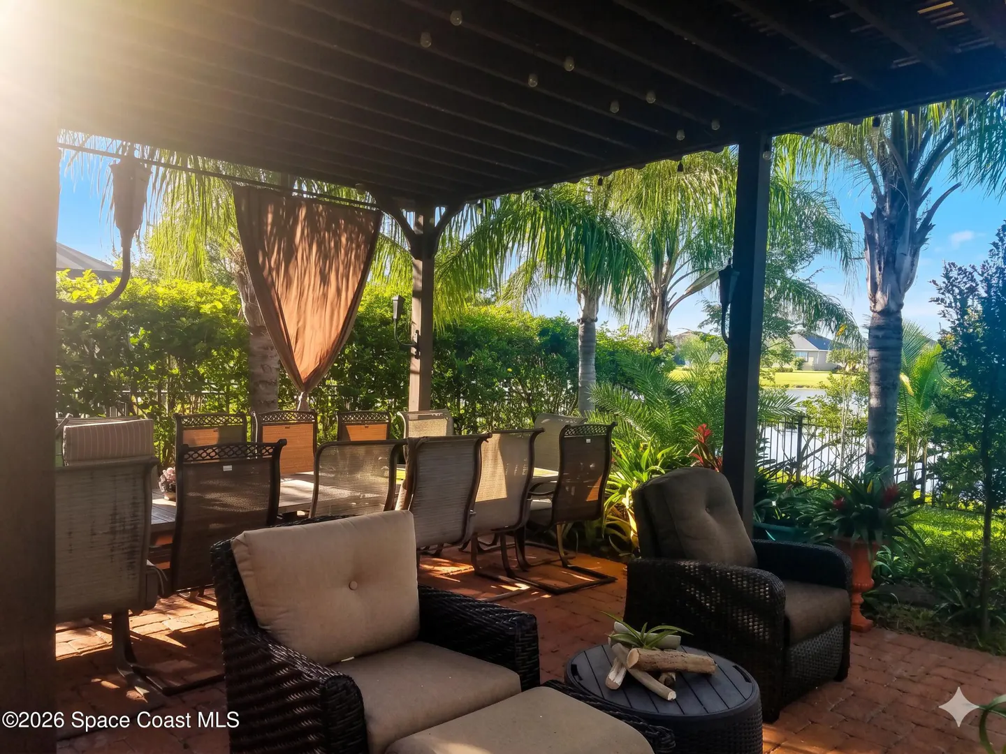 Outdoor patio with wicker furniture, a brown pergola, and lush green landscaping. A lake is visible in the background.