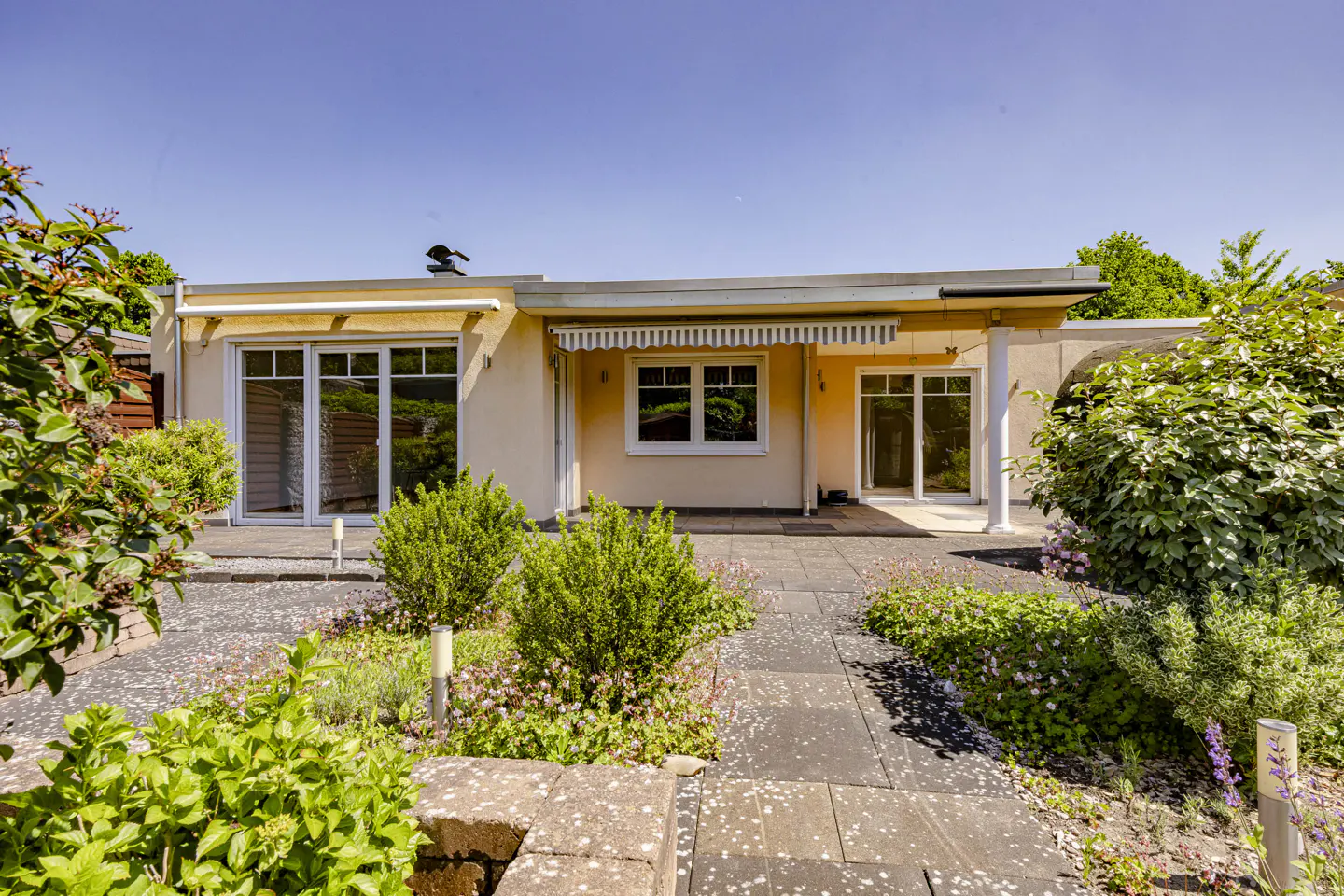 A single-story beige house with a patio, glass doors, and a striped awning, surrounded by a lush garden on a sunny day.