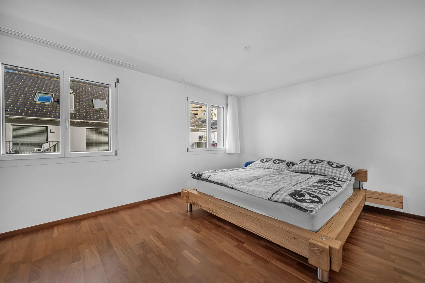 Bright bedroom with hardwood floors, white walls, and a wooden platform bed with patterned bedding. Two windows offer a view of neighboring houses.