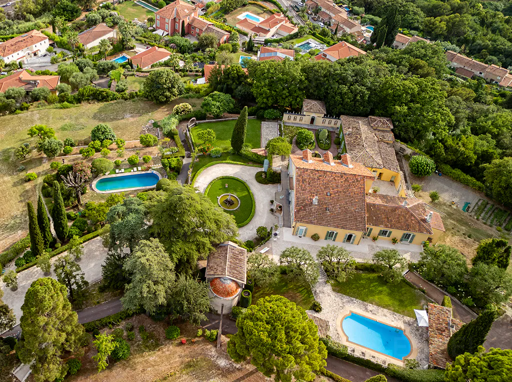 Aerial view of a large yellow house with a red tile roof, surrounded by green trees and lawns, with two blue swimming pools.