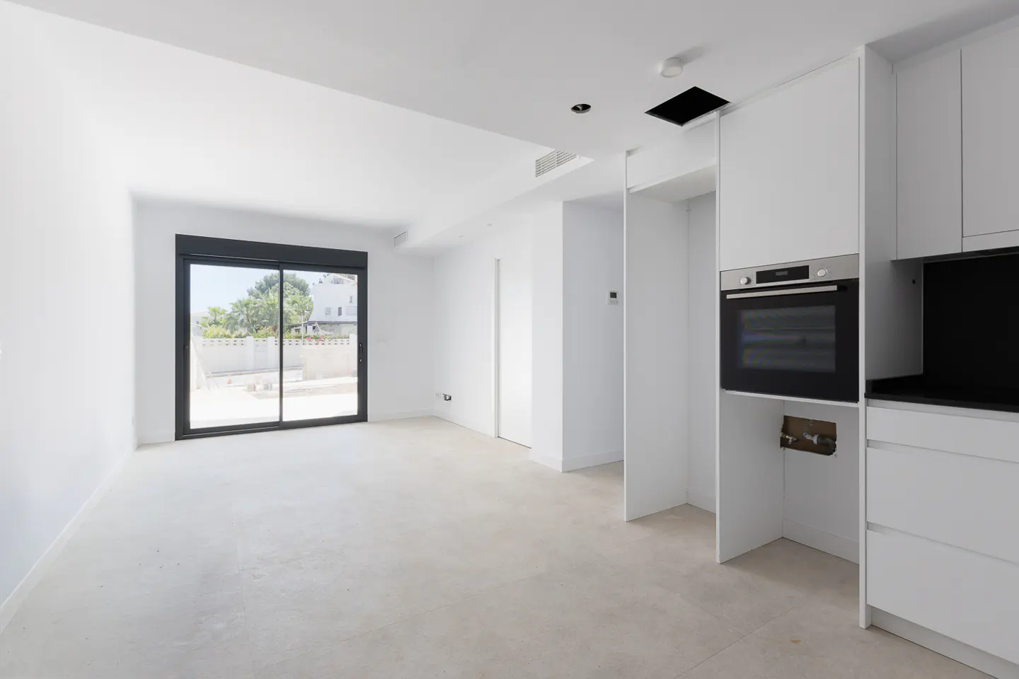 Modern white kitchen and living area with a built-in oven, light tile floors, and a sliding glass door to a patio.