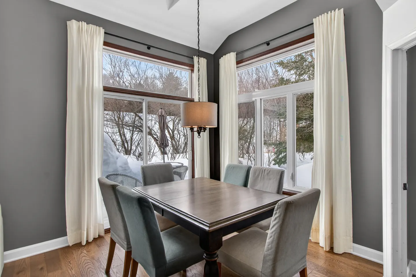 Dining room with gray walls, hardwood floors, and a square table with six chairs. Large windows with white curtains overlook a snowy landscape.