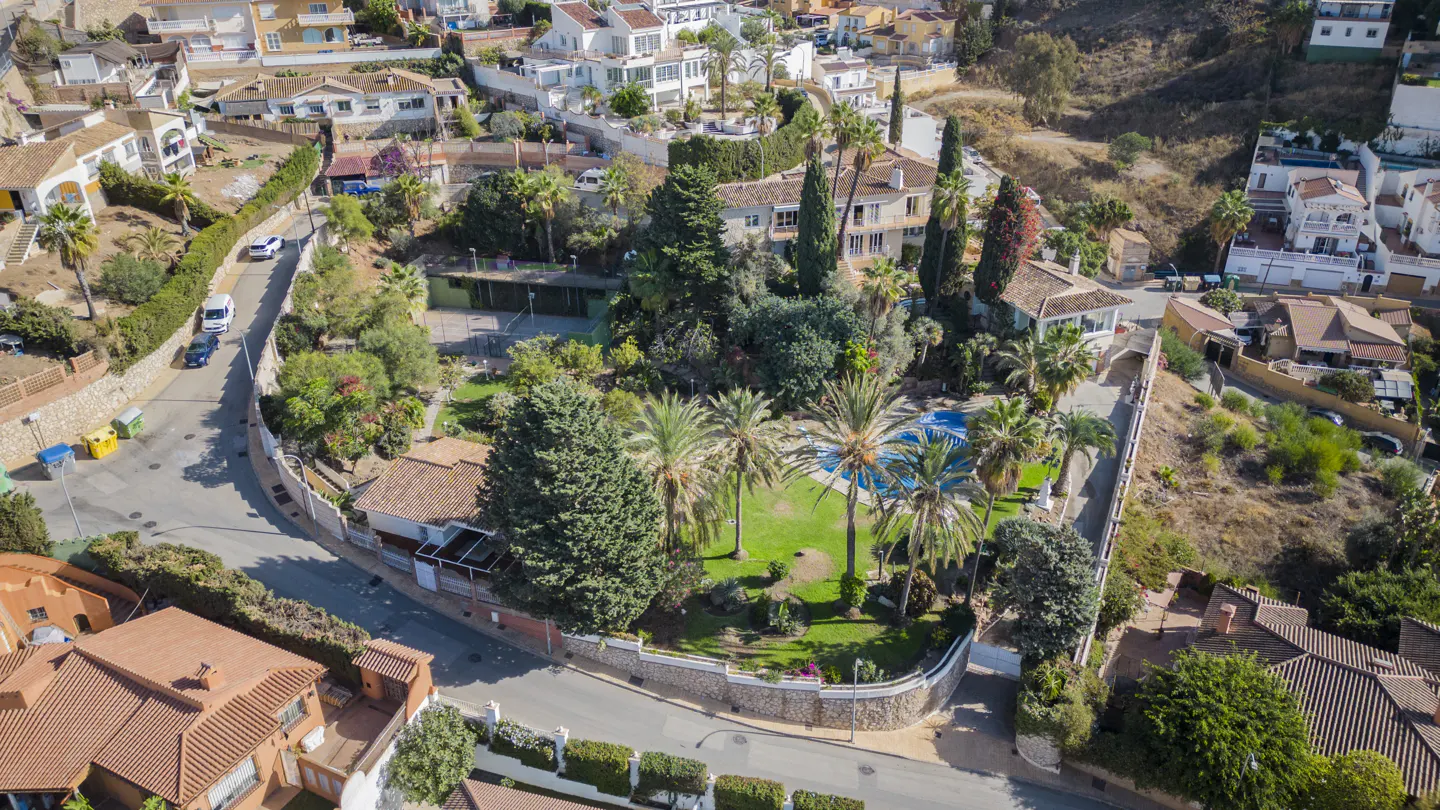 Aerial view of a Spanish villa with a pool, tennis court, and lush green gardens surrounded by white houses.
