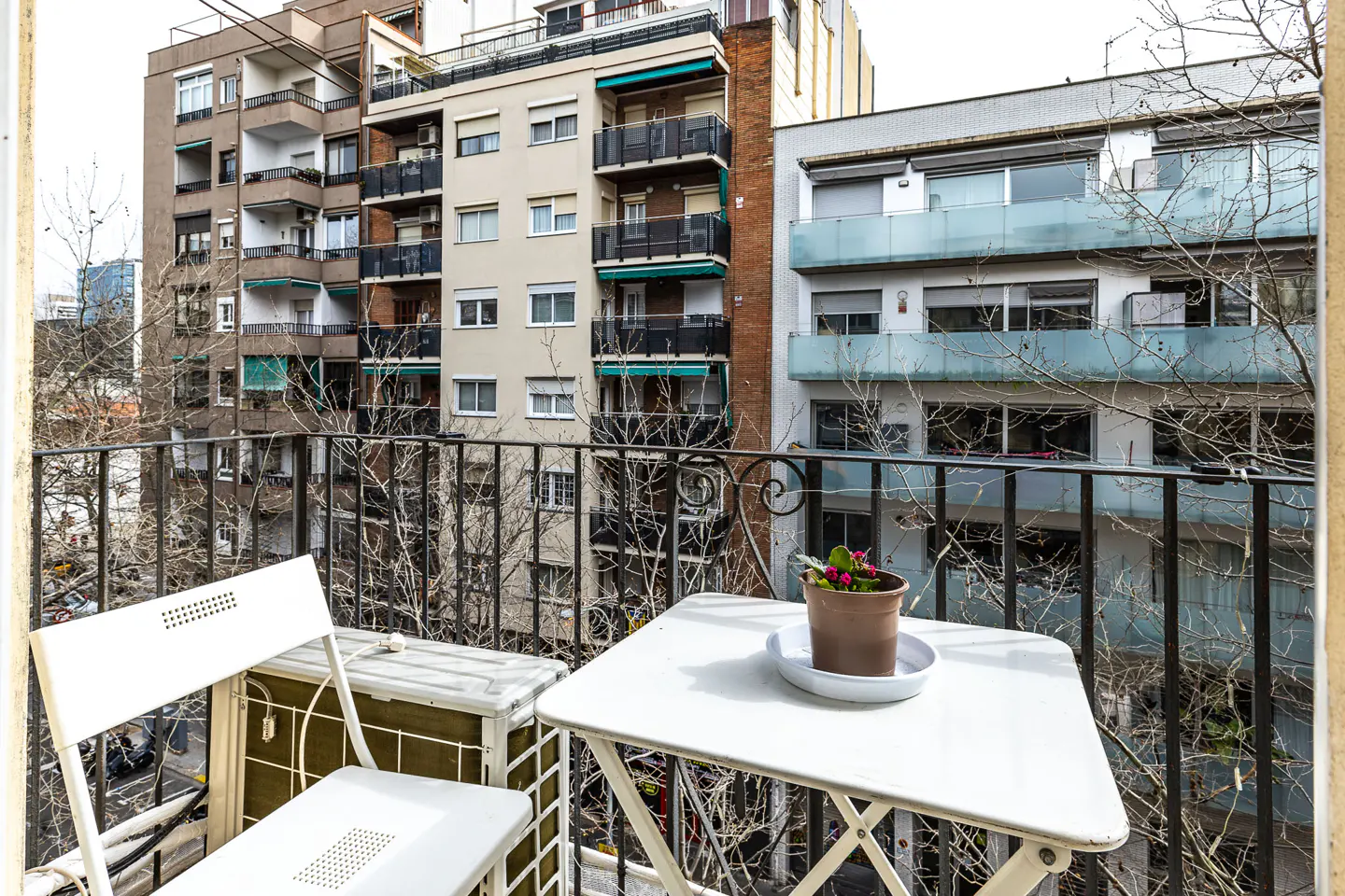 Balcony view with white table, chair, and potted flowers. Buildings are visible in the background.