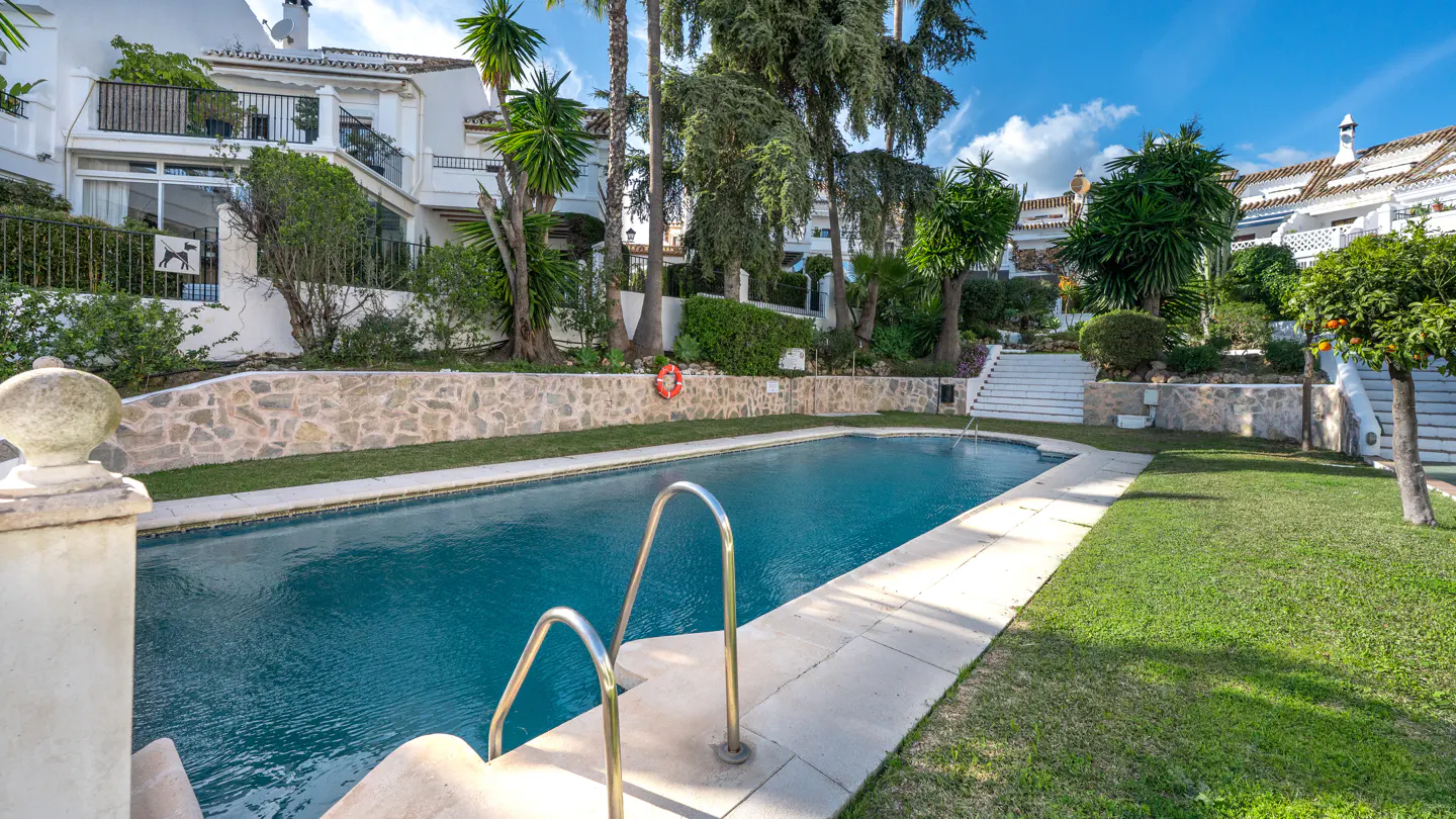 Outdoor pool with metal ladder, surrounded by green grass and stone walls. White buildings and trees in the background.