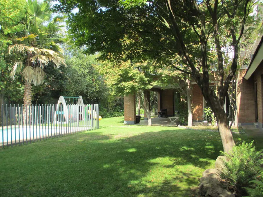 Lush green lawn with trees, a playground, and a brick house with a covered patio. A white fence borders a pool.