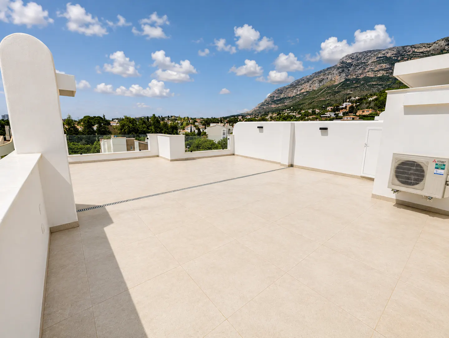 Wide rooftop terrace with beige tiles, white walls, and mountain views under a blue sky with clouds.