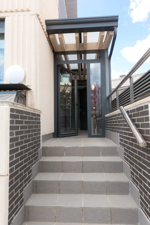 Exterior view of a building entrance with gray stone steps, a glass door under a wooden pergola, and brick walls.