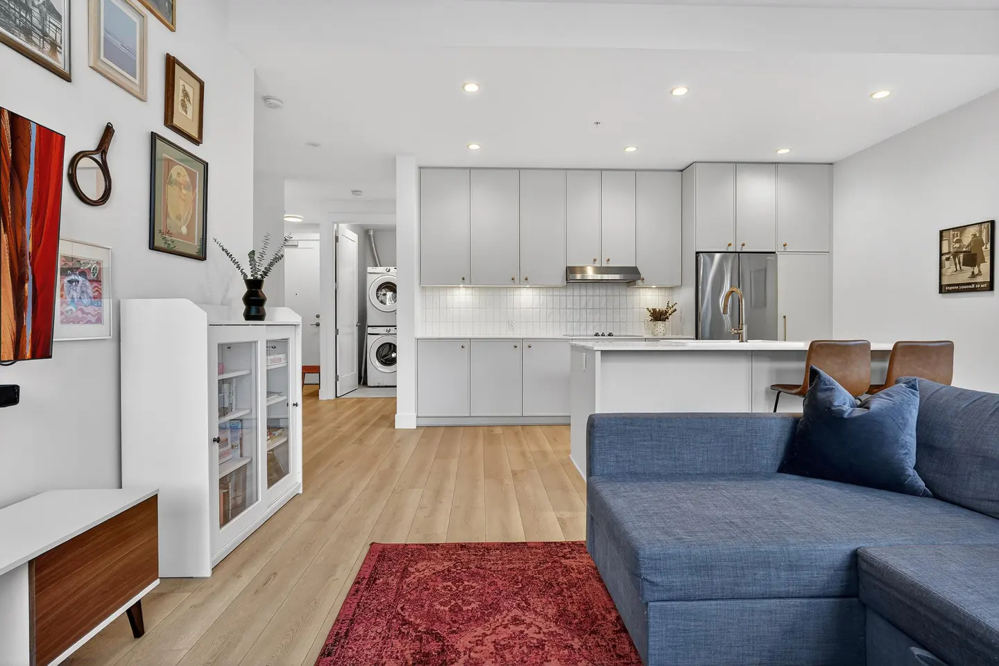 Open-concept living space with a blue sofa, wood floors, and a gray kitchen. Washer and dryer are visible in the background.