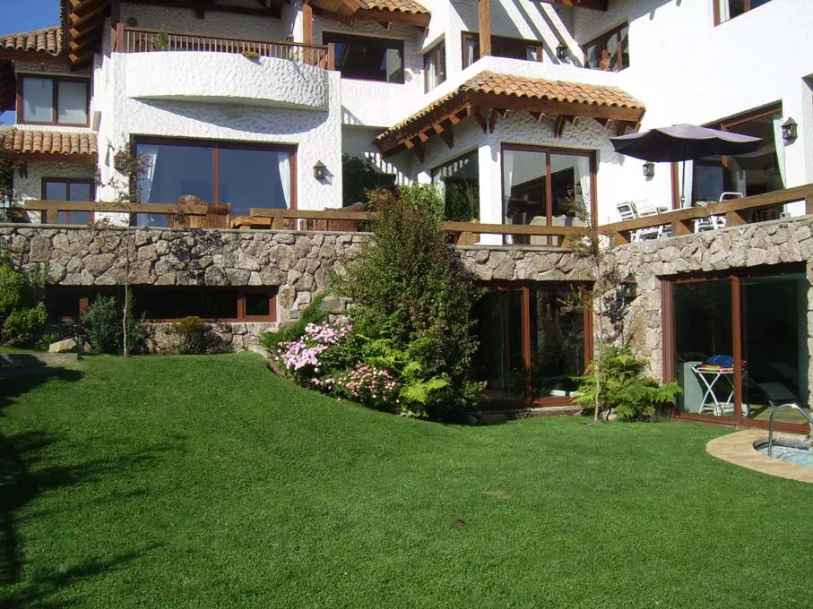 Multi-level white house with stone accents, brown trim, and a green lawn. A patio with an umbrella is visible.