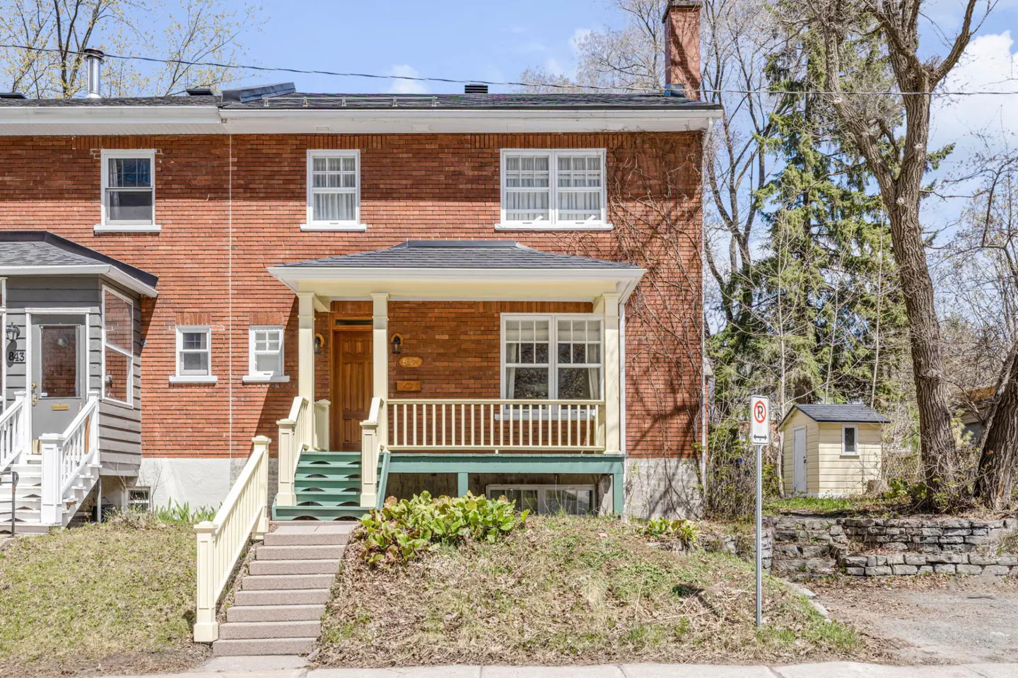 Red brick two-story house with a porch, white trim, and a green staircase leading to the front door. A small shed is visible in the background.