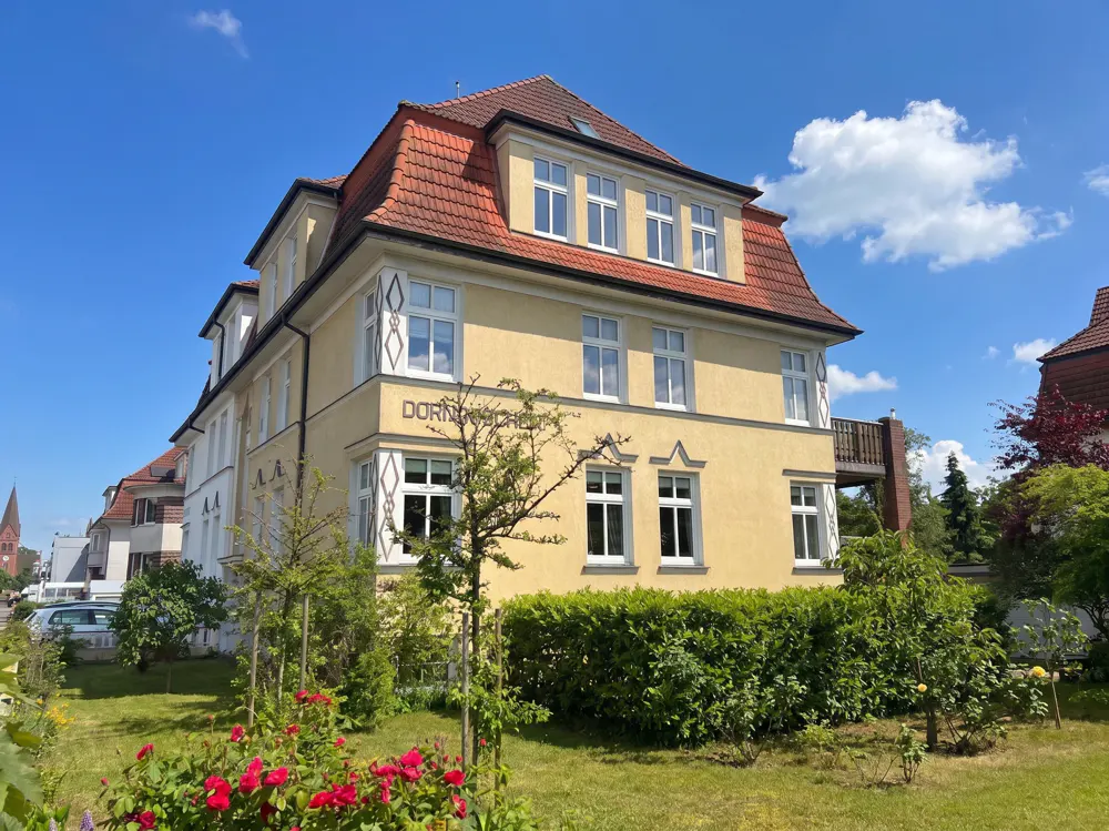 A three-story yellow apartment building with a red tile roof, surrounded by a green lawn, bushes, and red rose bushes.