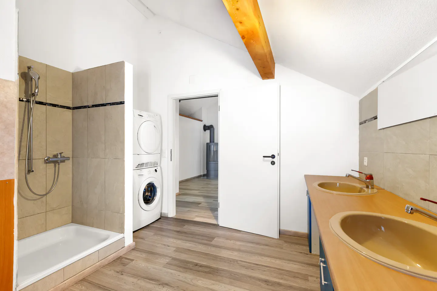 Bright bathroom with wood-look floors, white walls, and a wood beam. Features a shower, stacked washer/dryer, and double sink vanity.
