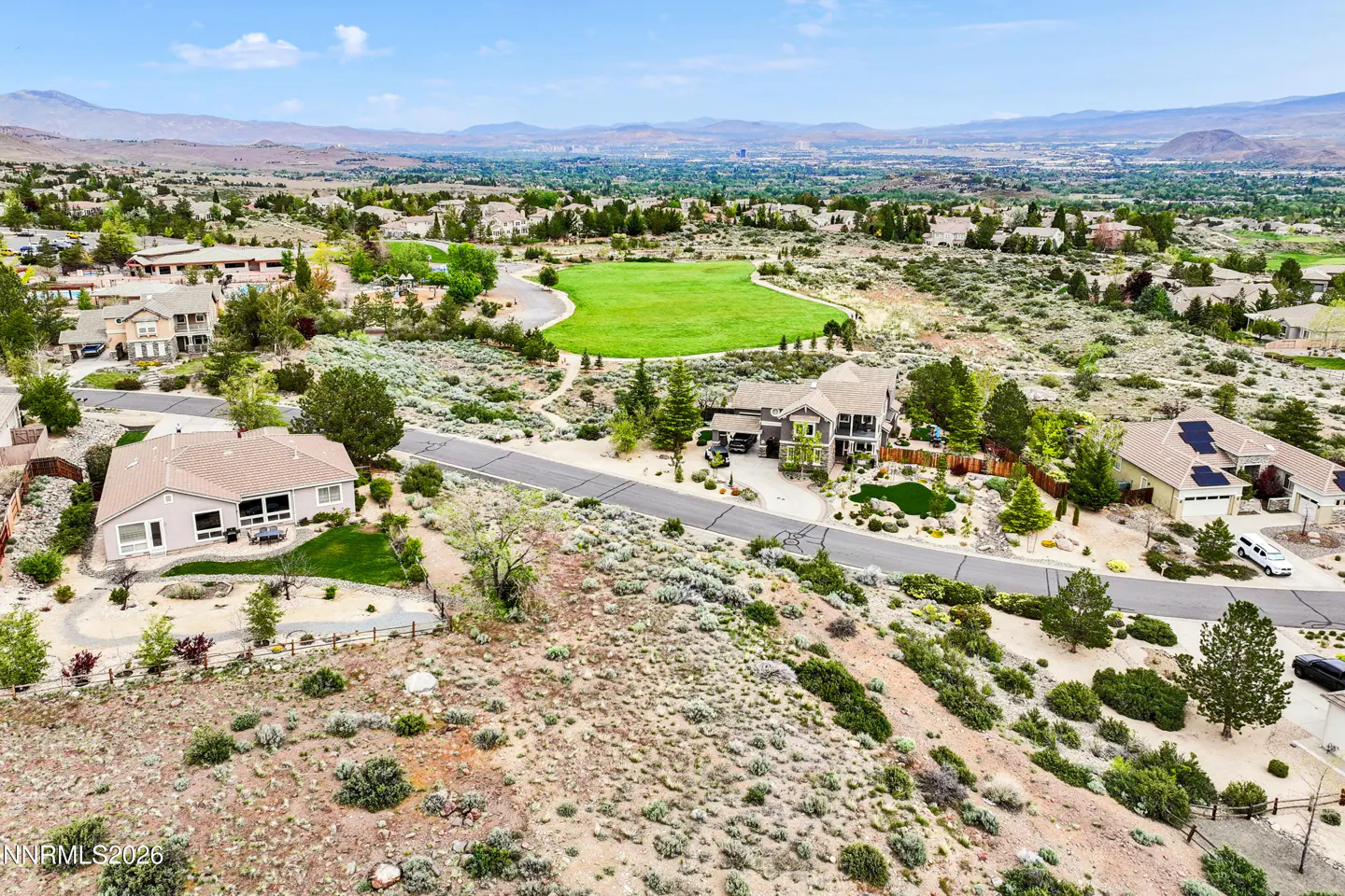 Aerial view of a residential area with houses, a green field, and mountains in the background under a blue sky.