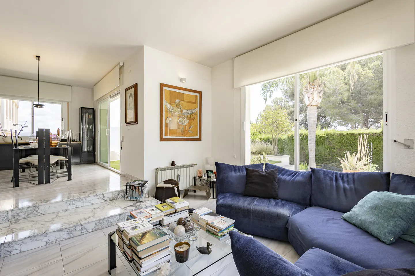 Bright living room with a blue sectional sofa, glass coffee table stacked with books, and a view of a garden through windows.