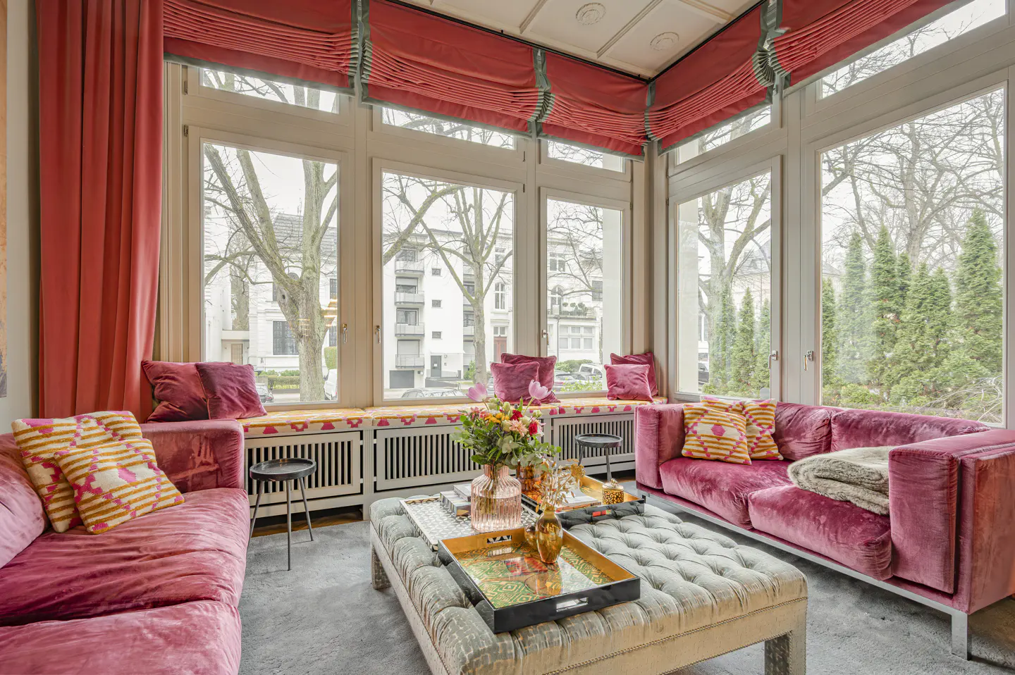 Bright sunroom with pink velvet sofas, patterned pillows, and a tufted ottoman. Large windows overlook trees and buildings. Red curtains and blinds add warmth.