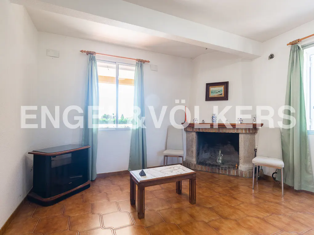 Living room with brown tile floor, fireplace, two chairs, a dark wood TV stand, and a wood coffee table. Light blue curtains hang by the windows.