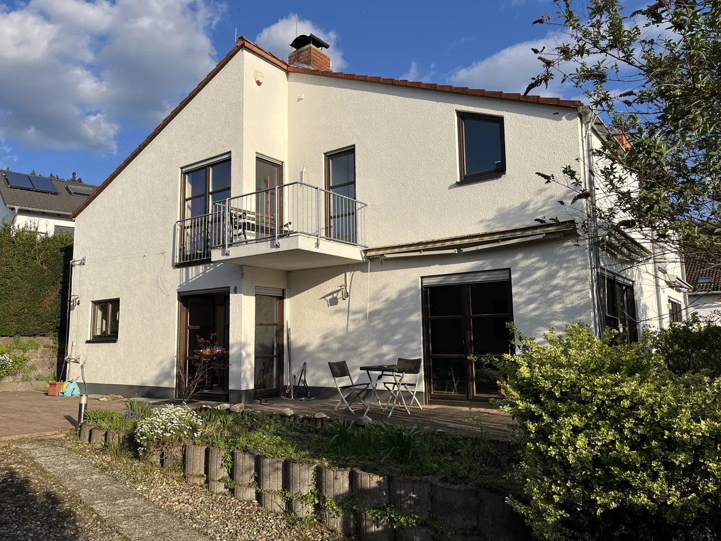 Two-story white house with a balcony, dark-framed windows, and a brick chimney under a blue sky. A patio with chairs sits in front.