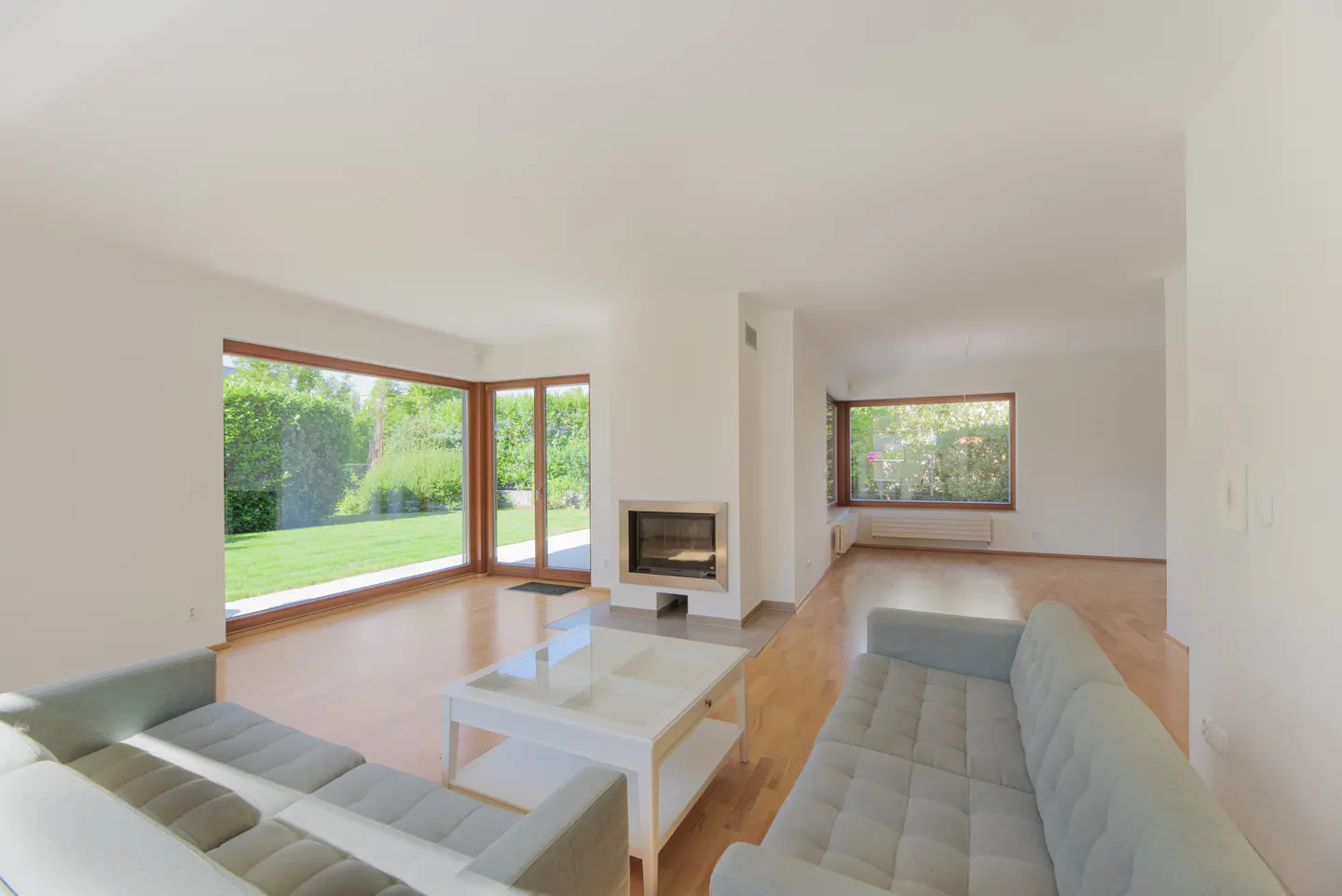 Bright living room with hardwood floors, a gray sofa, and a white coffee table. A fireplace is centered between large windows overlooking a green lawn.