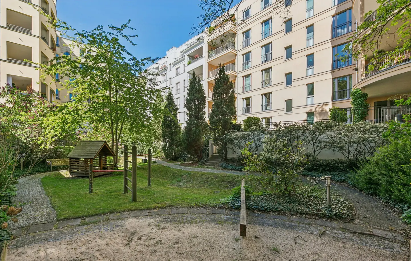 Apartment complex courtyard with a playground, green grass, trees, and a sand pit on a sunny day.