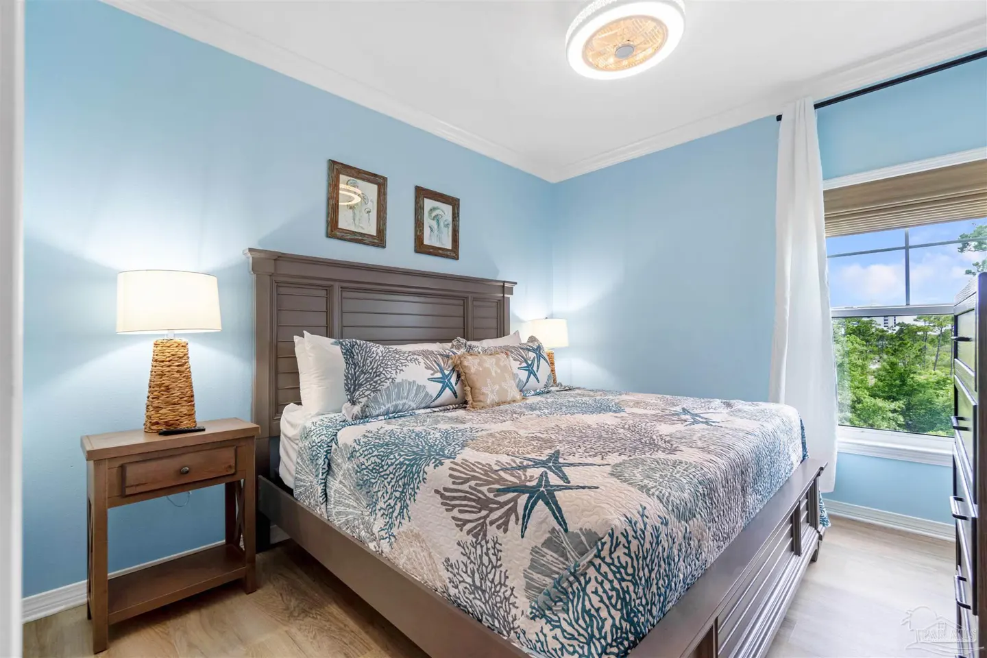 Bedroom with blue walls, a queen bed with a starfish patterned quilt, and a nightstand with a lamp. A window shows trees outside.