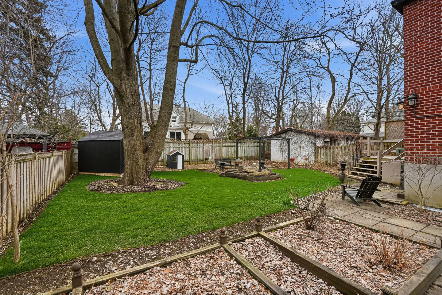 A backyard with green grass, a large tree, a black shed, a wooden fence, and a brick house.
