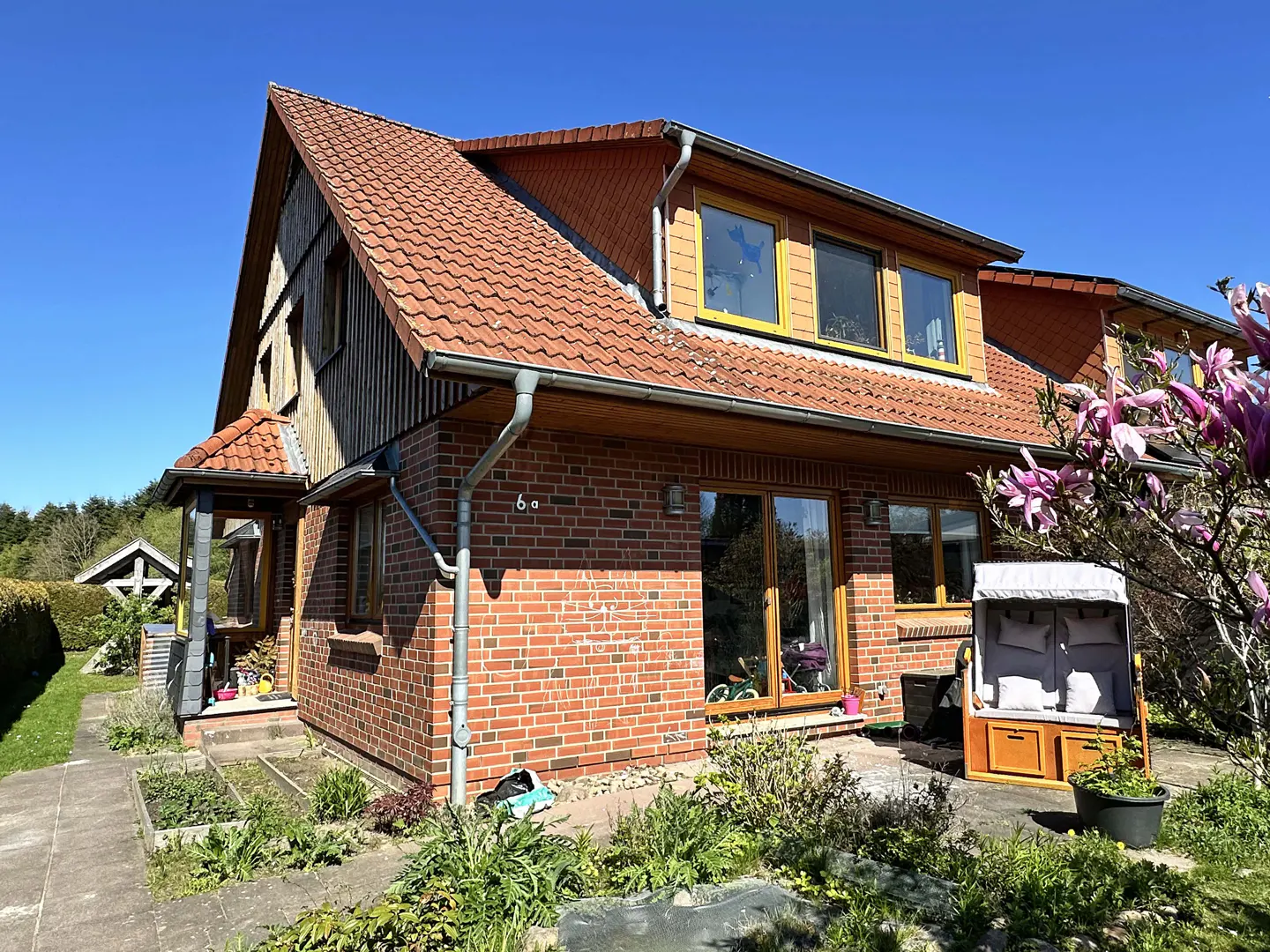 Two-story brick house with a red tile roof and wooden accents, set against a clear blue sky. A beach chair sits on the patio.