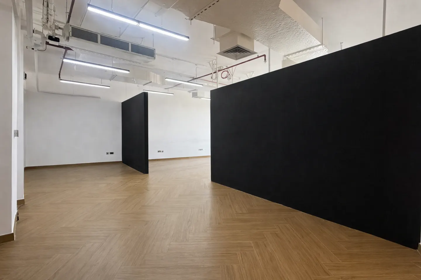 An empty office space with herringbone wood floors, white walls, and black partitions. Overhead lighting and ductwork are visible.