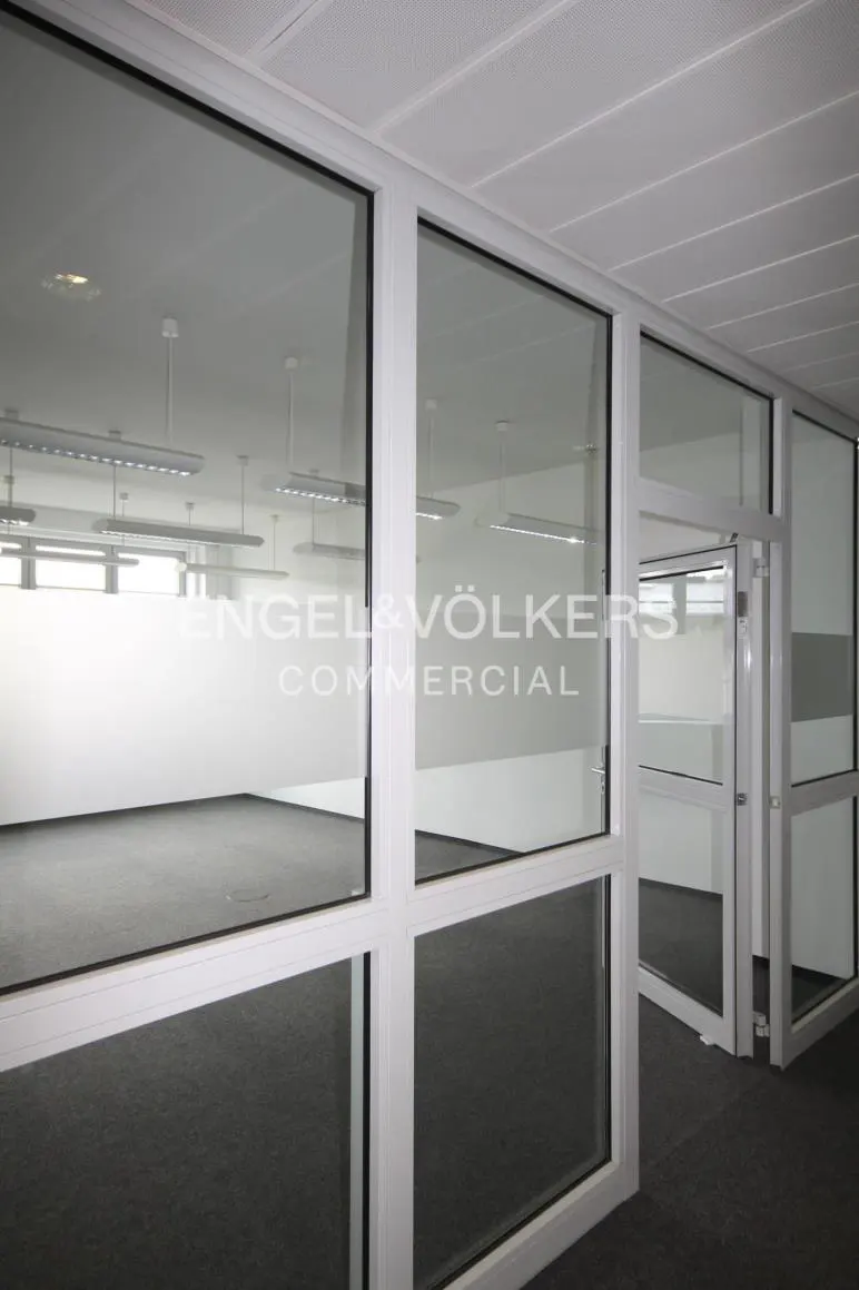 Office space with white walls, gray carpet, and large windows. Engel & Völkers Commercial logo on glass.