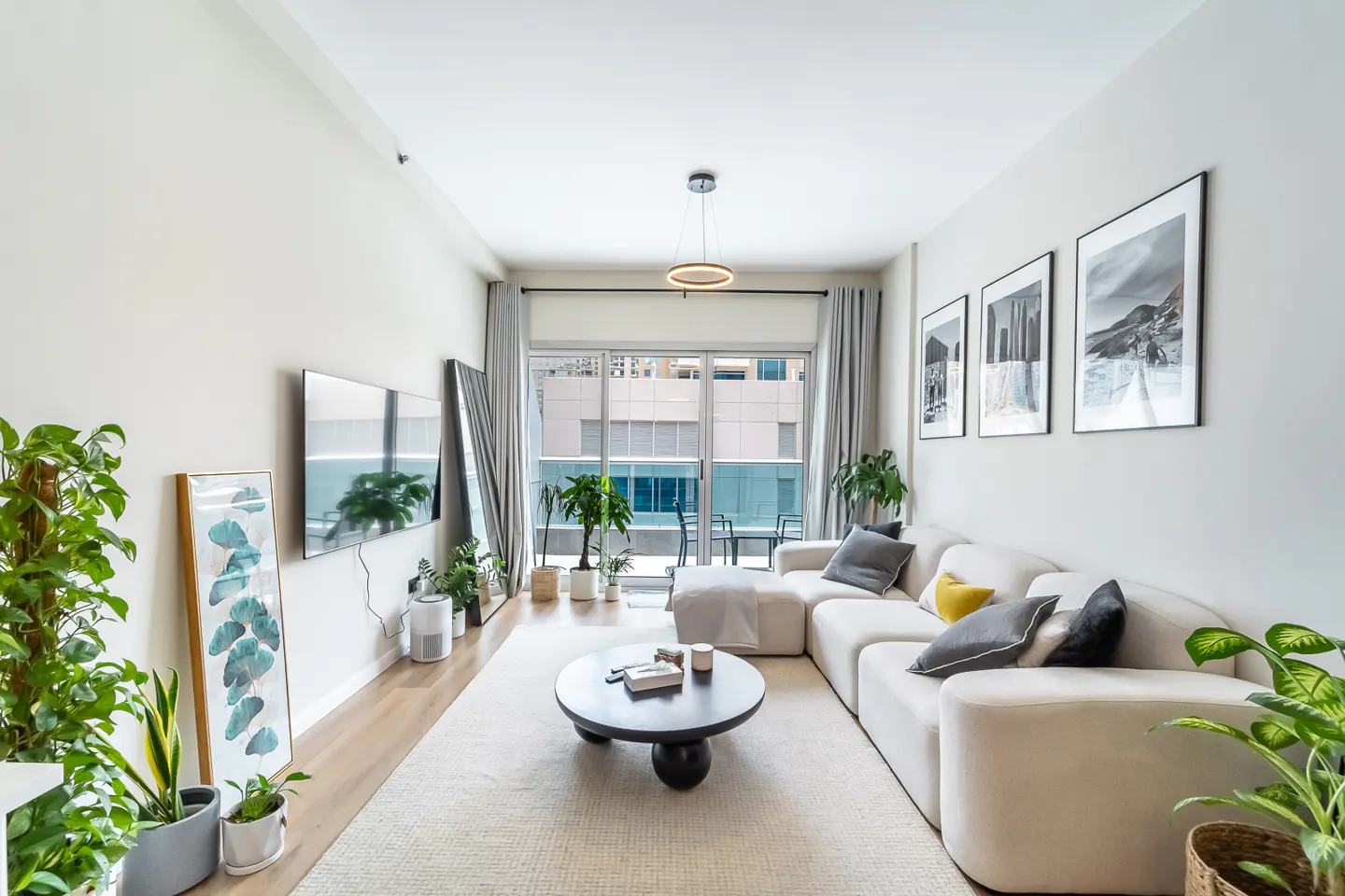 Bright living room with a white sectional sofa, black coffee table, and potted plants. A TV and framed art decorate the walls.