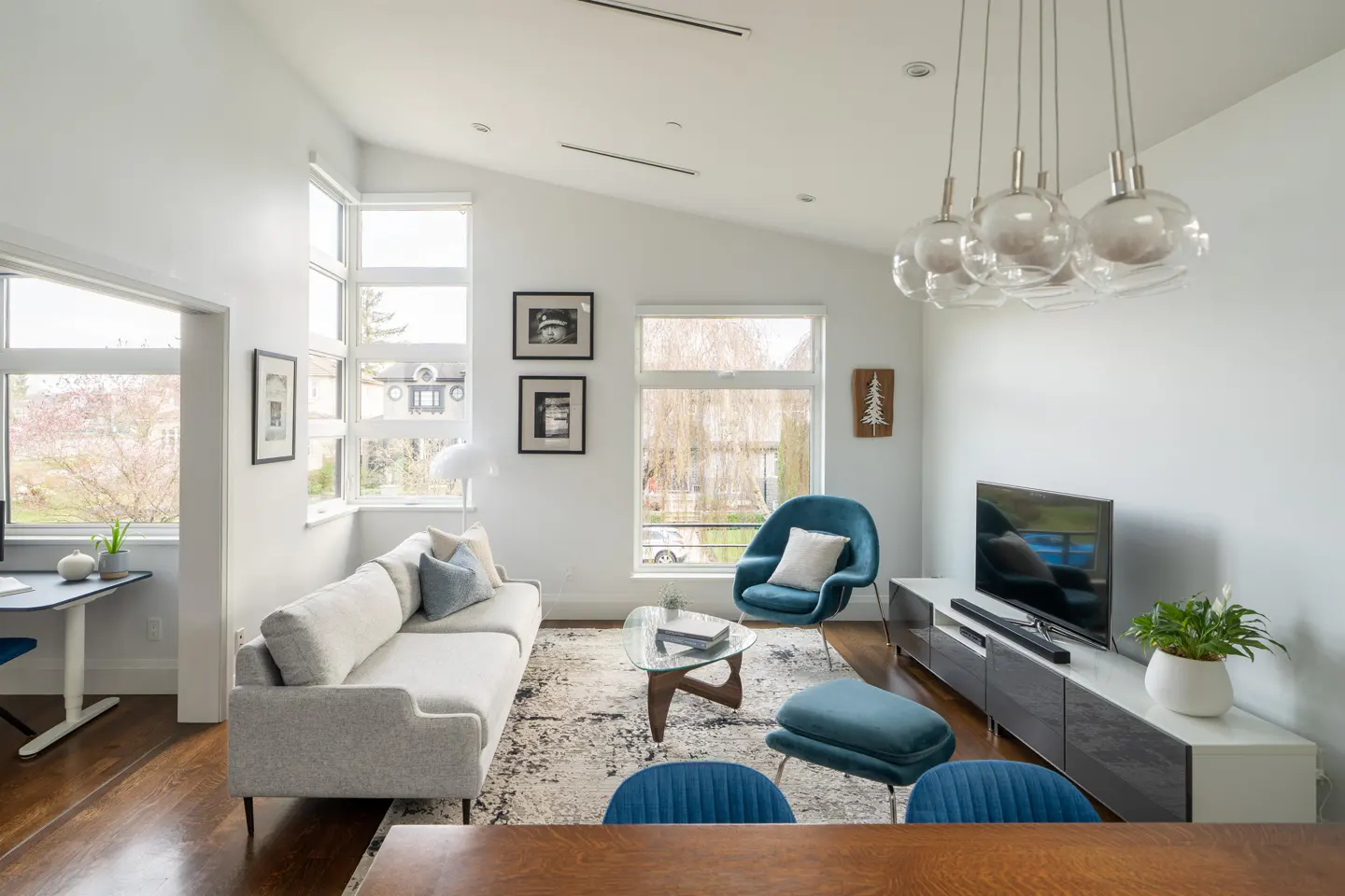 Bright living room with white walls, wood floors, and large windows. A gray sofa, blue chair, and glass coffee table sit on a patterned rug. A TV and modern lighting complete the space.