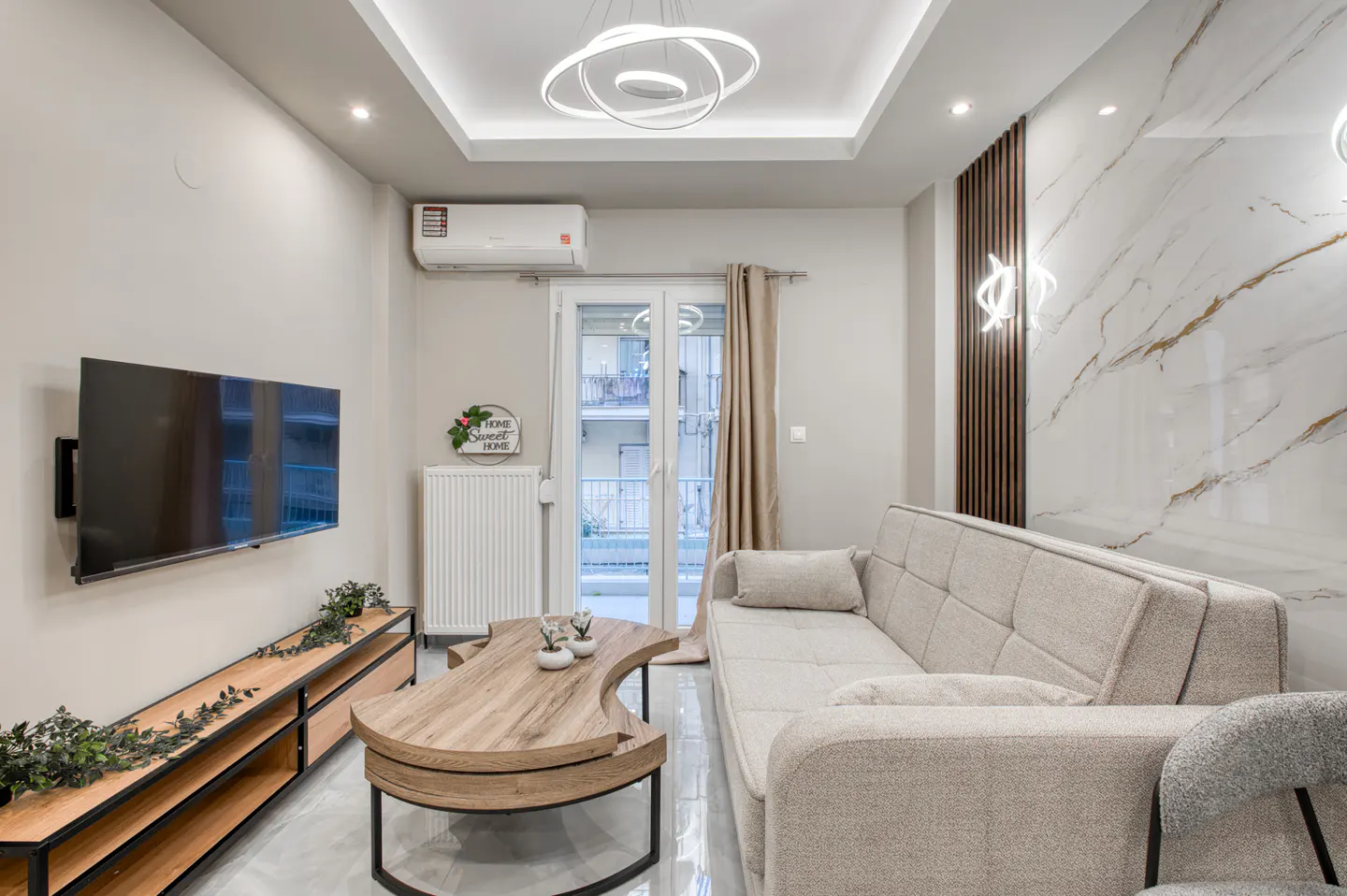 Living room with a beige sofa, wood coffee table, TV, and marble accent wall. Modern lighting fixtures illuminate the space.