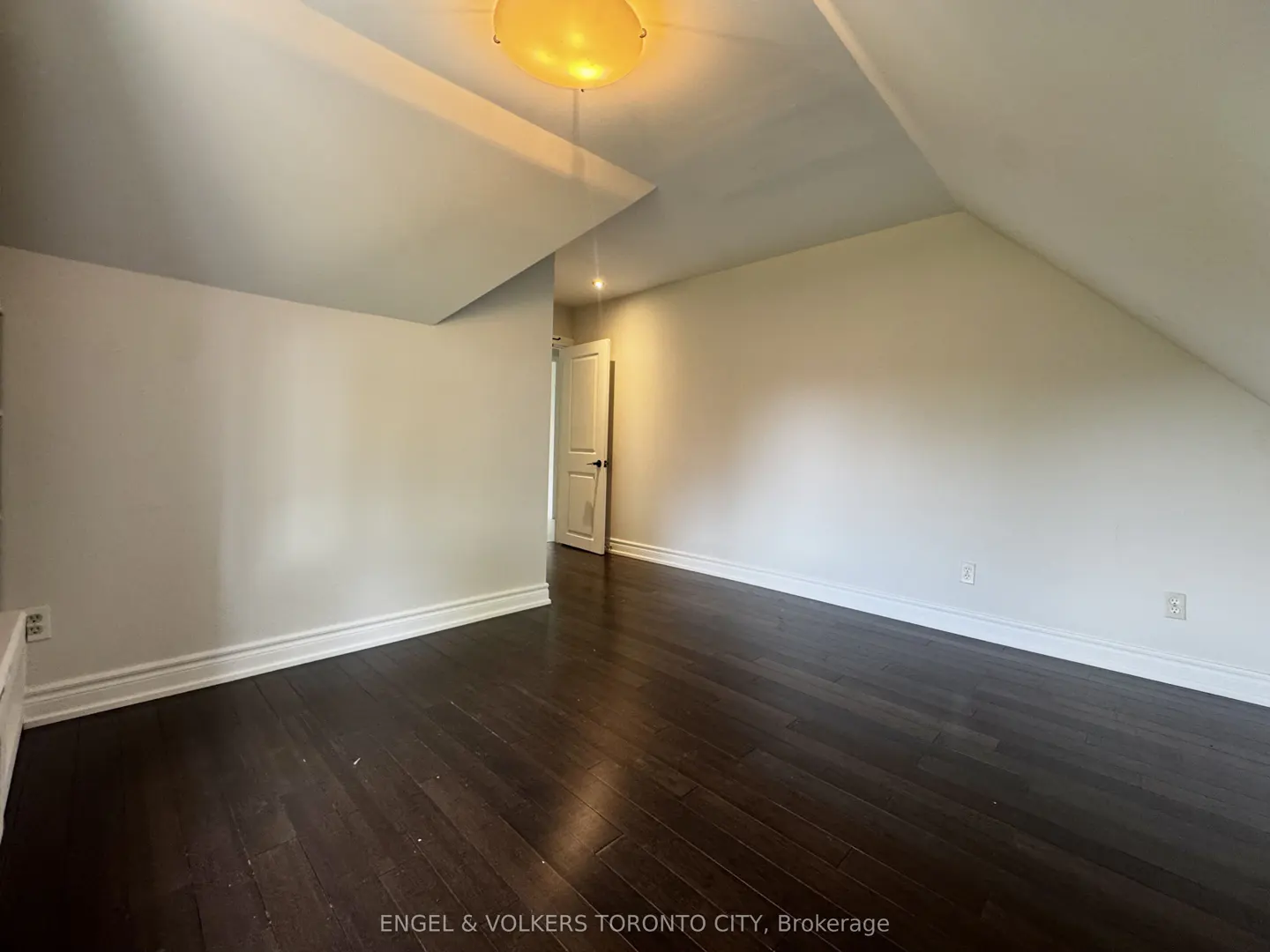 Attic room with dark wood floors, white walls, and a sloped ceiling. A white door is visible in the background. A round light fixture is on the ceiling.