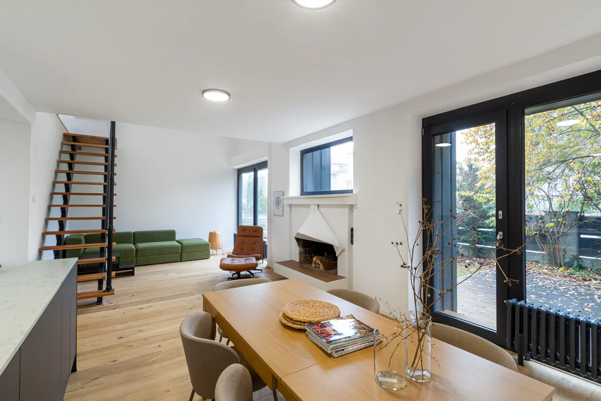 Bright, modern living room with wood floors, a dining table, fireplace, green sofa, and a staircase. Large windows offer a view of the outdoors.