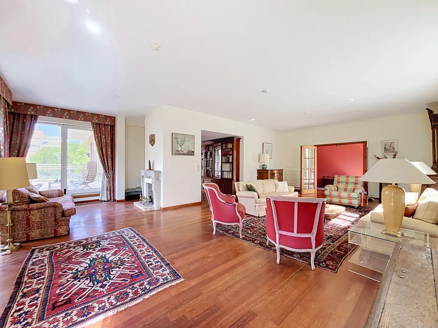Spacious living room with hardwood floors, red and cream armchairs, patterned rugs, and a fireplace near a balcony door.