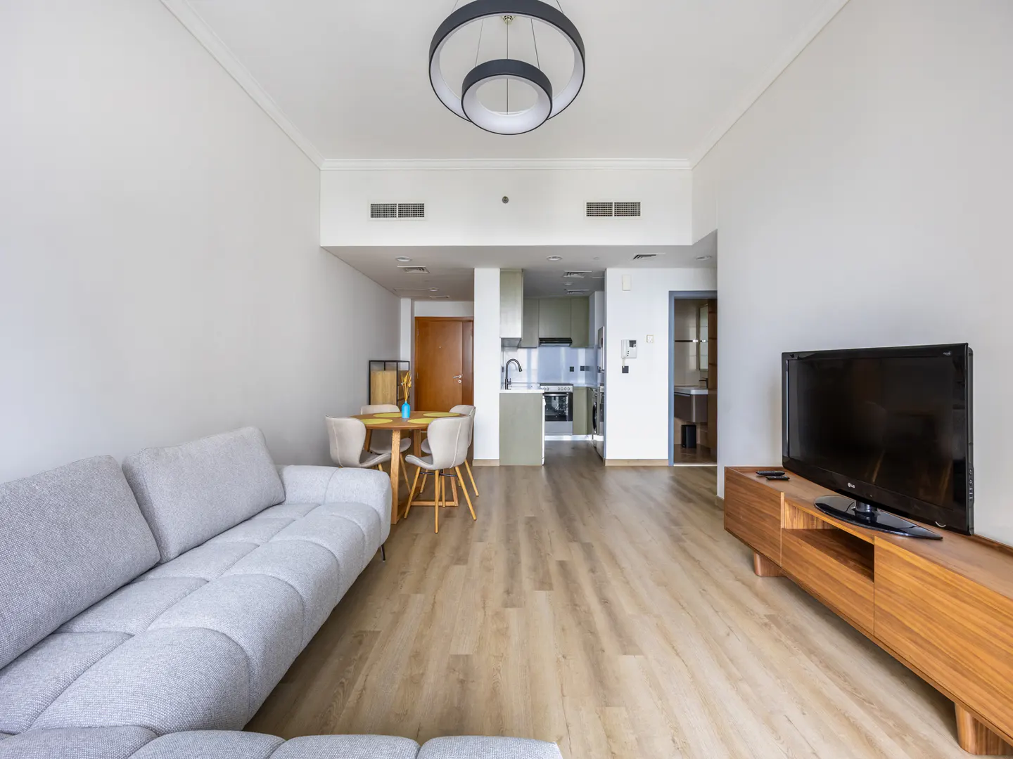 A bright, modern apartment interior with a gray sofa, dining table, TV, and open kitchen.