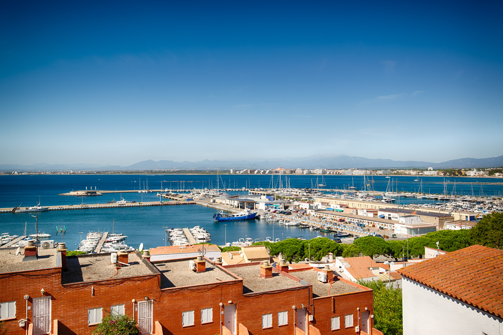 High-angle view of a sunny coastal town with red-tiled roofs overlooking a harbor filled with boats under a clear blue sky. [AI-generated text]