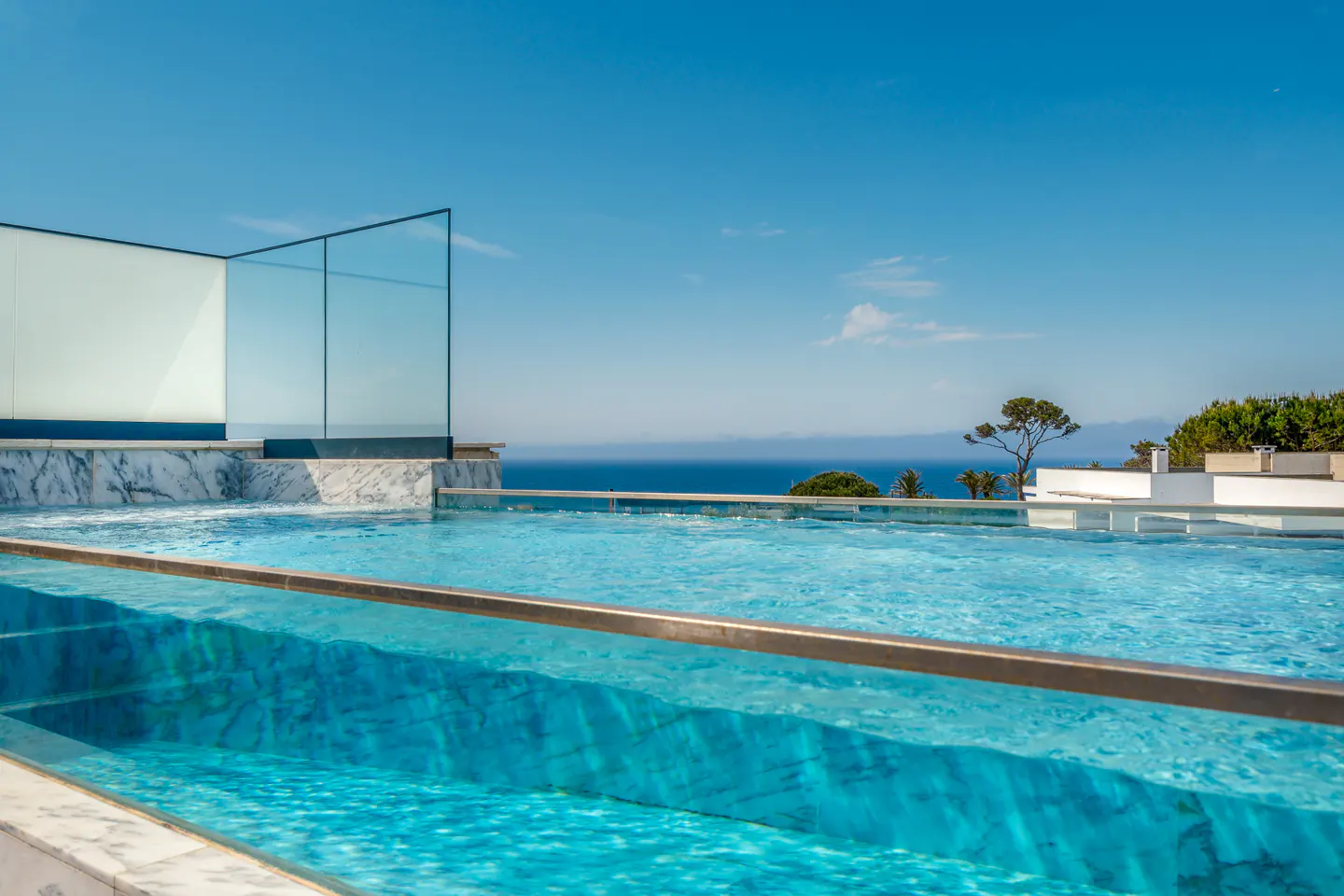 Infinity pool with clear glass edge, marble trim, and ocean view under a blue sky.
