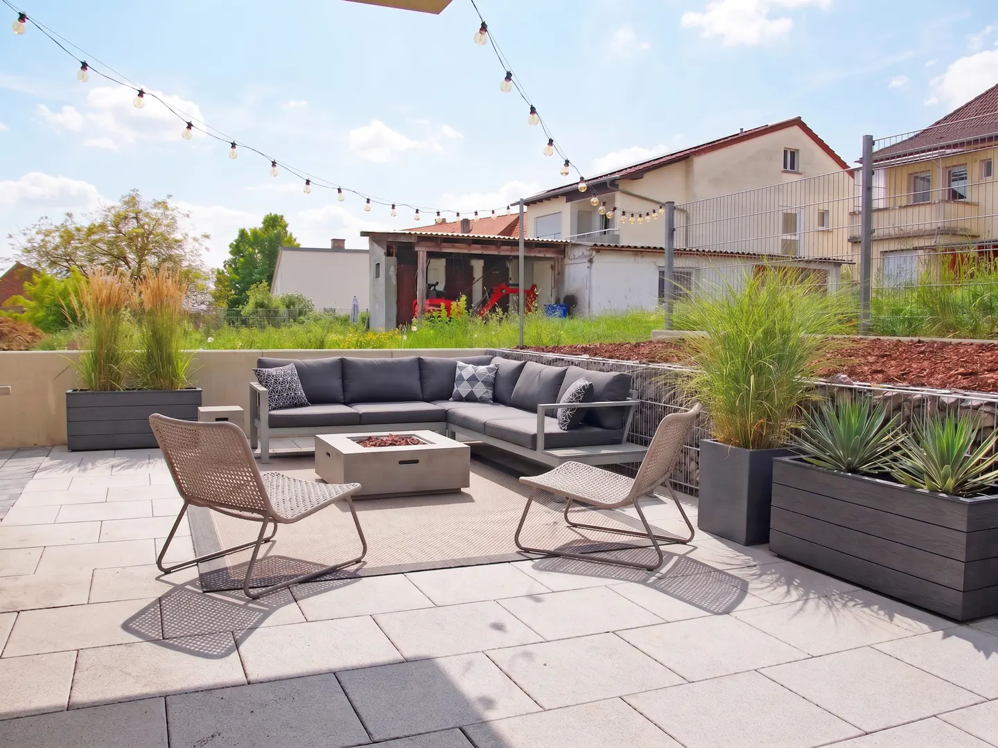 Outdoor patio with gray sectional sofa, fire pit, and two chairs on a stone patio. String lights hang overhead.