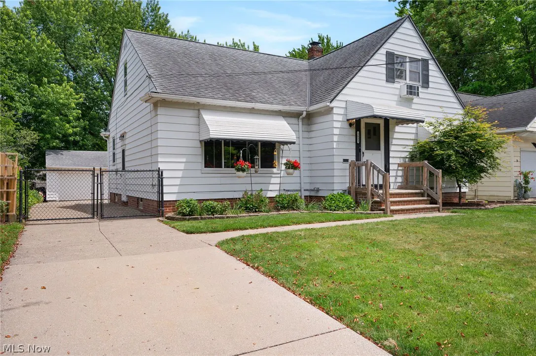 White cape cod house with a concrete driveway, front lawn, and a chain-link fence leading to a detached garage.