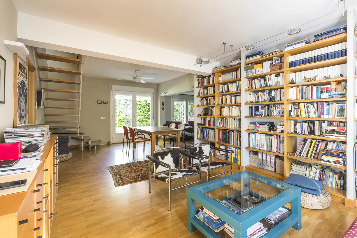 Bright living room with hardwood floors, filled bookshelves, a blue coffee table, and cowhide chairs. A dining table is visible in the background.