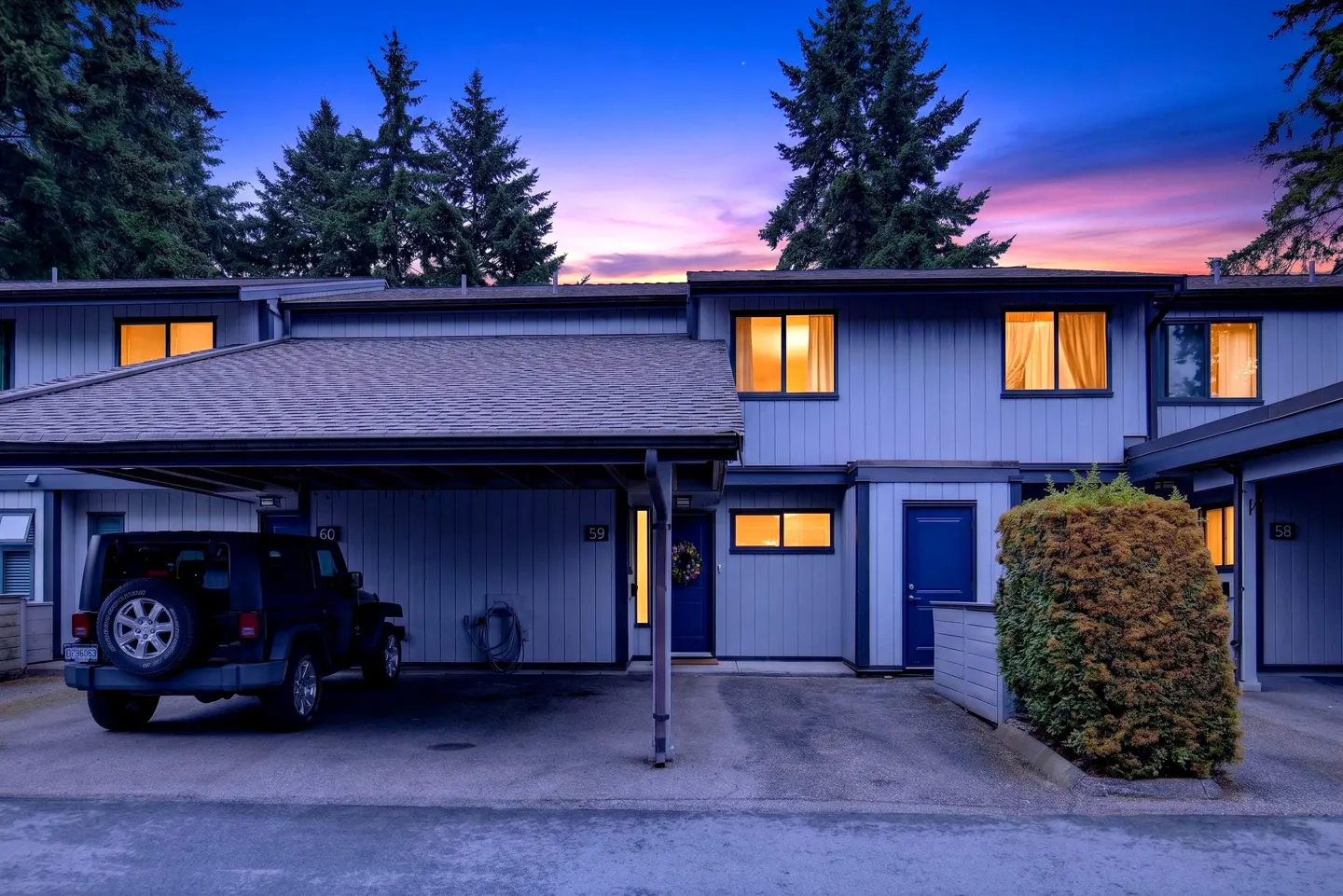 Blue townhouse exterior at dusk with a carport, a parked black SUV, and a vibrant purple and orange sunset sky.