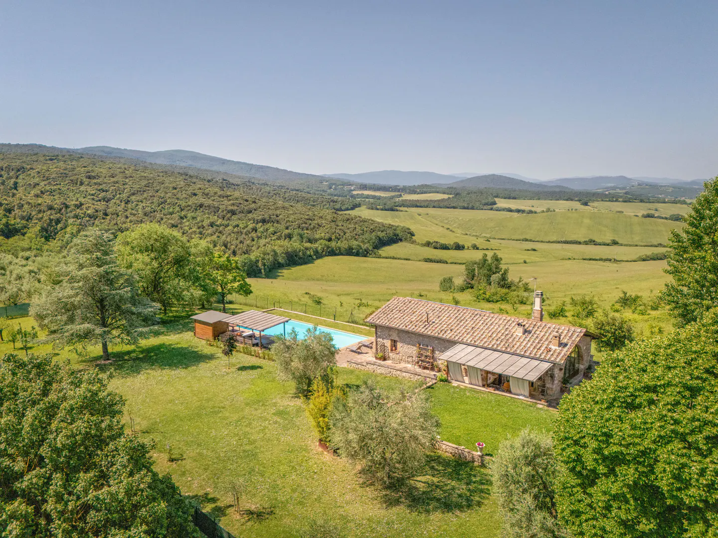 Aerial view of a stone farmhouse with a tiled roof, swimming pool, and patio set in a lush, rolling Tuscan landscape.