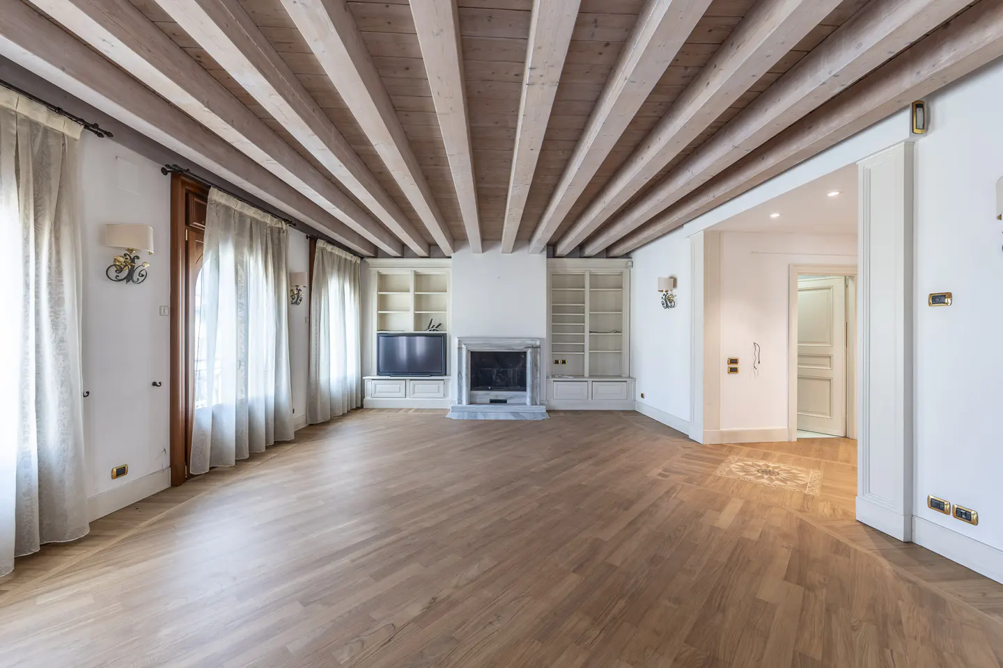 Bright, empty living room with wood floors, beamed ceiling, and fireplace. White walls, curtains, and built-in shelving.