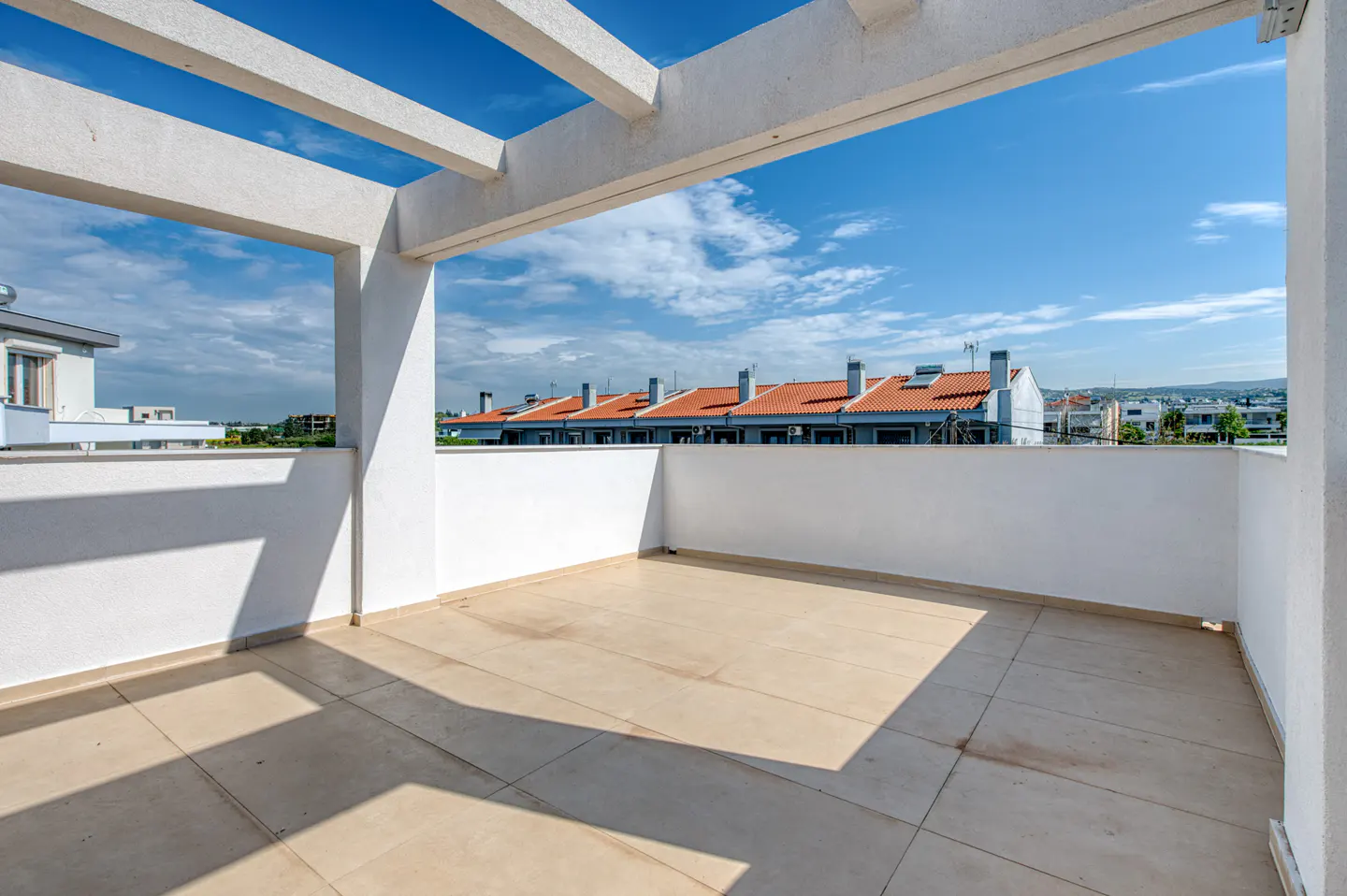 A bright, empty balcony with beige tile flooring, white walls, and a pergola roof overlooks a neighborhood with orange-roofed houses under a blue sky.