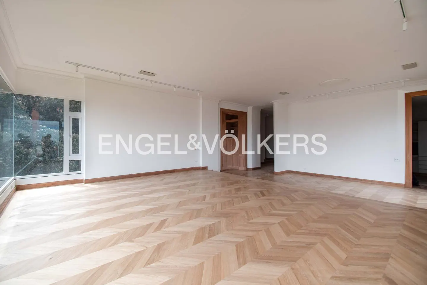Bright, empty room with herringbone wood floors, white walls, and large windows. Engel & Völkers logo is superimposed on the image.