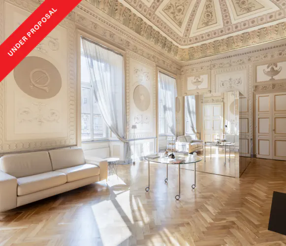 Ornate room with herringbone wood floors, cream walls, and a decorated ceiling. A cream sofa sits near a window with sheer curtains. "Under Proposal" banner.
